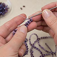 Close-up of hands using a steel crochet hook to create a beaded crochet necklace with purple thread and faceted beads.
